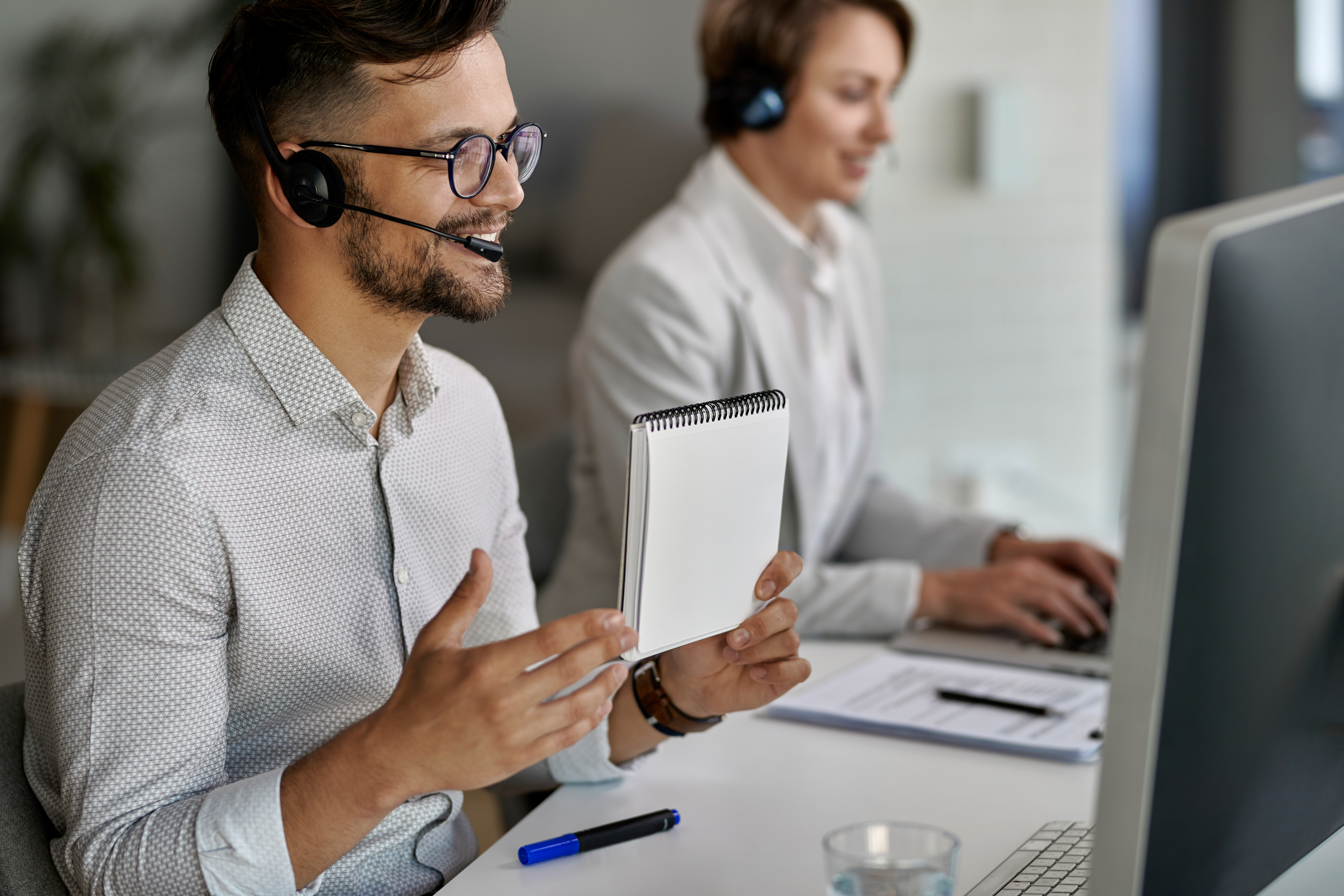A male customer service representative wearing a headset and glasses, holding a notepad, while another representative works on a computer in the background.